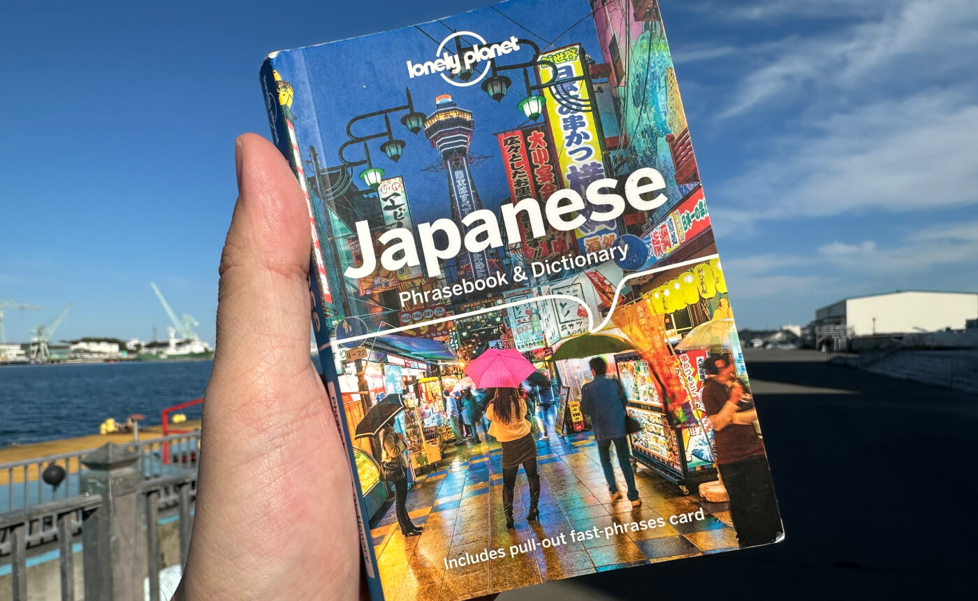 Hand håller i en liten bok med bild på en japansk gata med många lyktor och skyltar. Titeln lyder "Japanese Phrasebook & Dictionary".