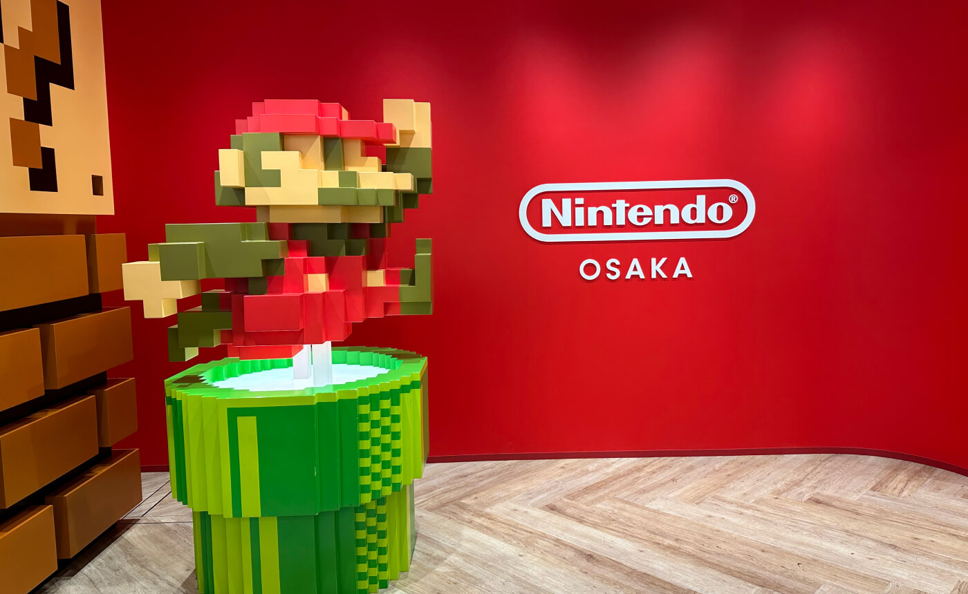 Bås vid ingen till butiken Nintendo Osaka. Mario hoppar upp ut ett rör. Skylten lyder "Nintendo Osaka".