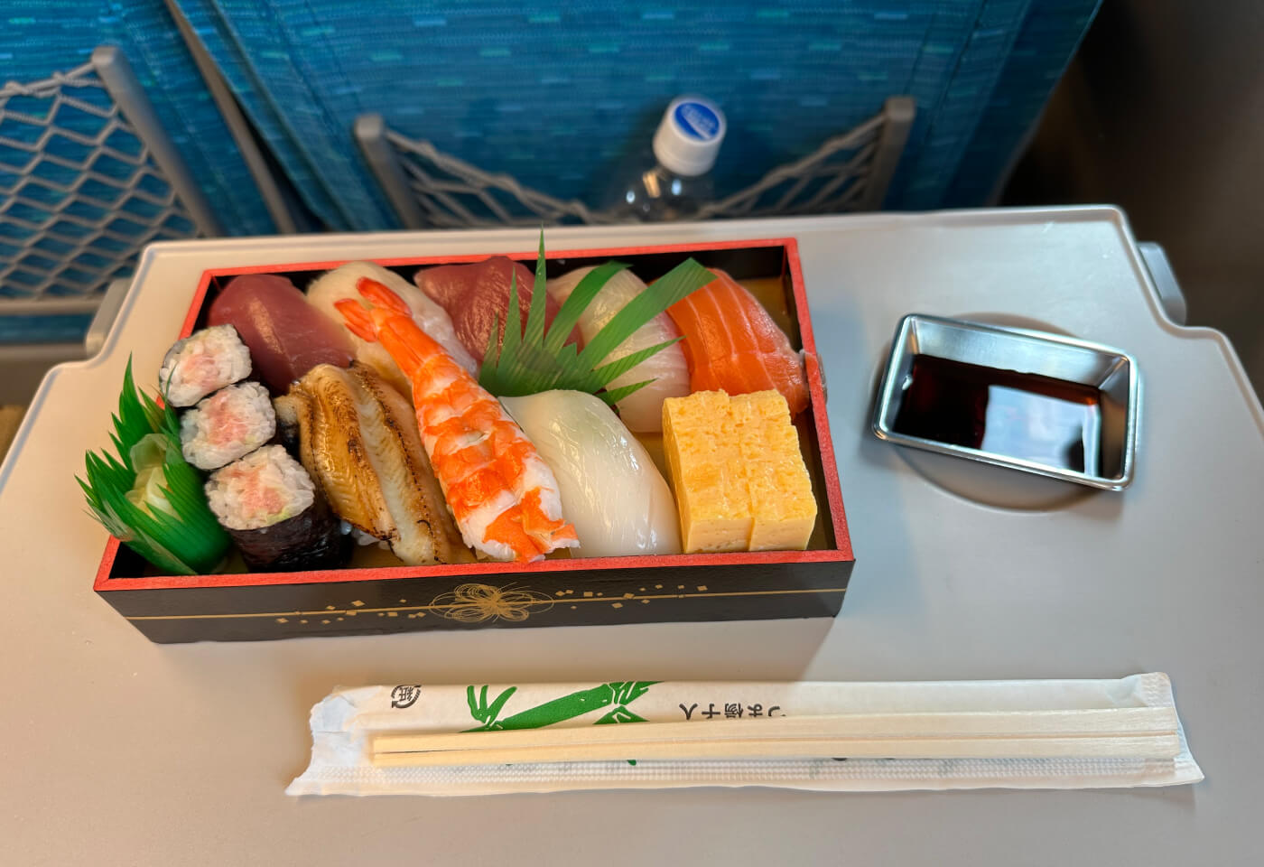 En fyrkantig låda med tretton bitar sushi på ett utfällbart bord på en tågplats. Till höger stör en liten skål med sojasås.