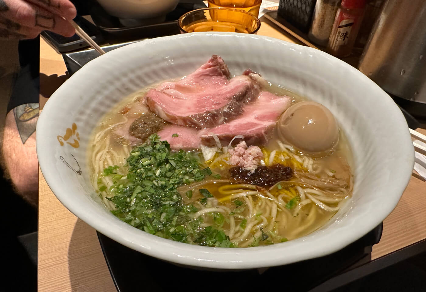 Vit skål med en stor portion ramen med ljus sky, ett kokt ägg, hackad lök, griskött mer mera.