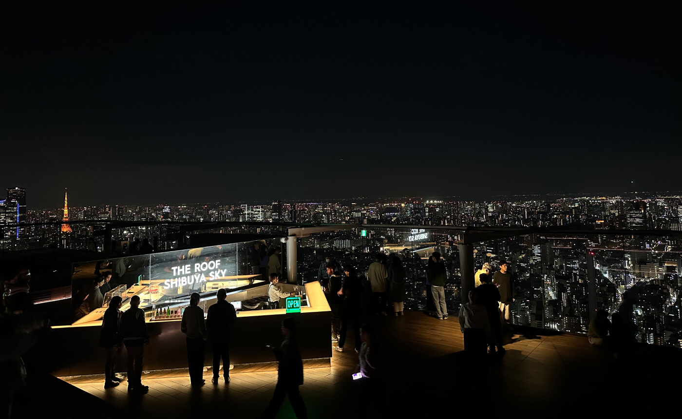 Takbar med en skylt som lyder "The Roof Shibuya Sky" på en öppen utkiksplats. I fjärran ser man en bred utsikt över Tokyo nattetid.