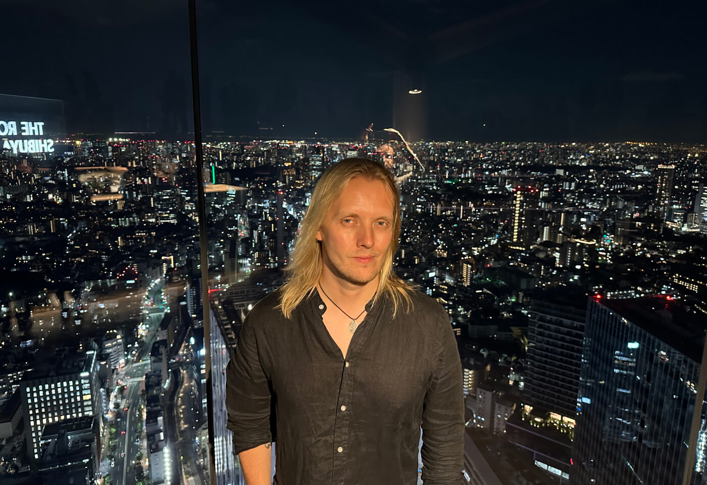 Jag står framför en glasskiva iförd mörk skjorta. Bakom glasskivan ser man en stor del av Tokyo. Det är mörkt ute.