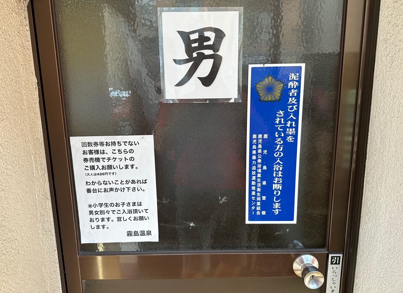 Mörk dörr med glas som har flera stora klistermärken med japansk skrift. Ett klistermärke har ett stort tecken för könet man.