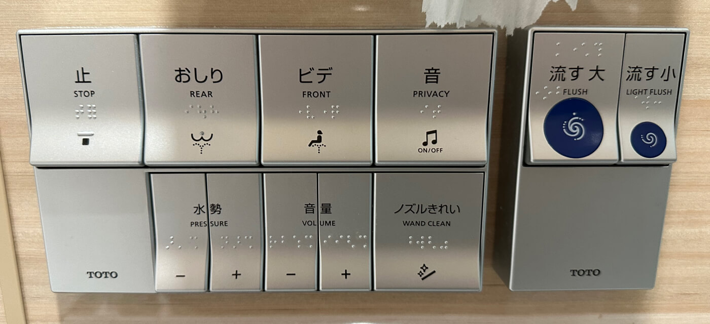 Ljusgrå väggdosa för att styra en japansk toalett. Alla elva knappar har text på både engelska och japanska. Vissa knappar har ikoner.