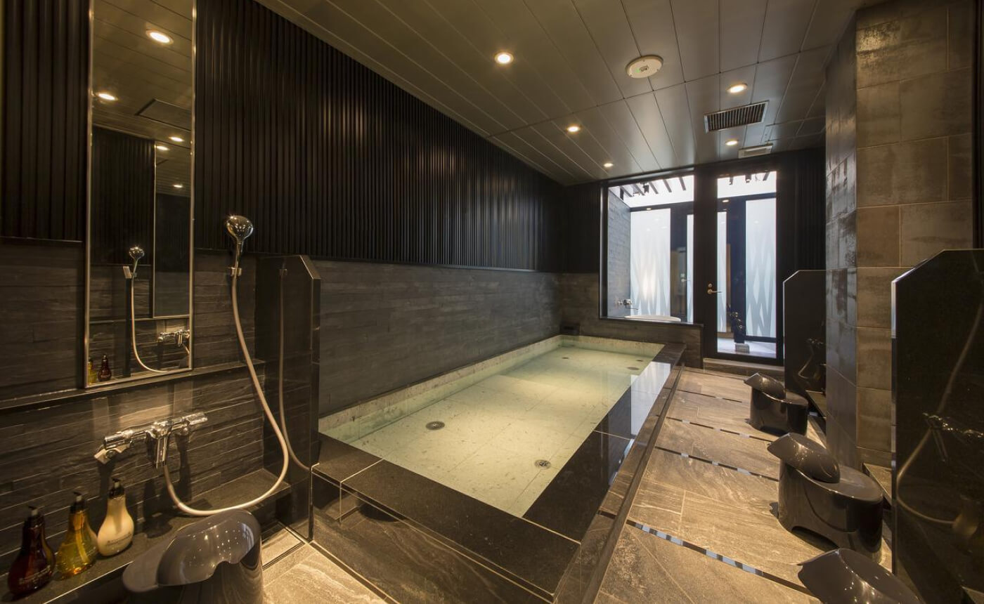 Pool omgiven av sittduschar i ett rim med mörka väggar. Längst bort är ett badkar utomhus.