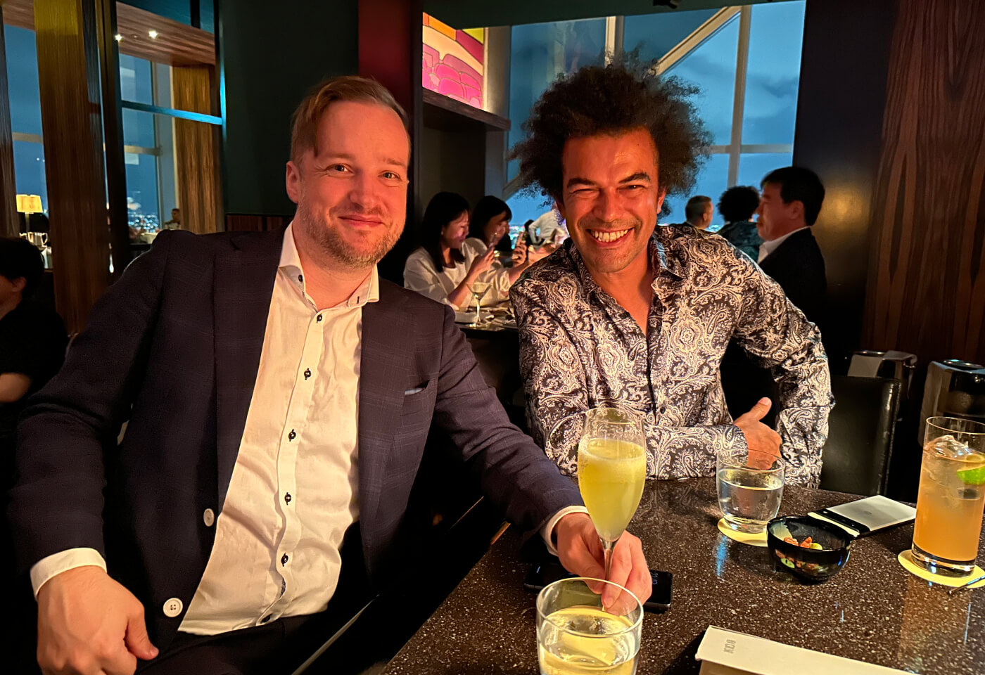 Dan Westin iförd mörk kostym tillsammans med Simon i ljus skjorta med svart mönster. De sitter vid en bar.