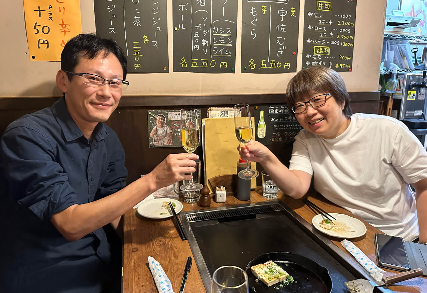 Ett japanskt par sitter vid ändan på ett bord med inbyggd stekyta. De skålar med varsitt vinglas.