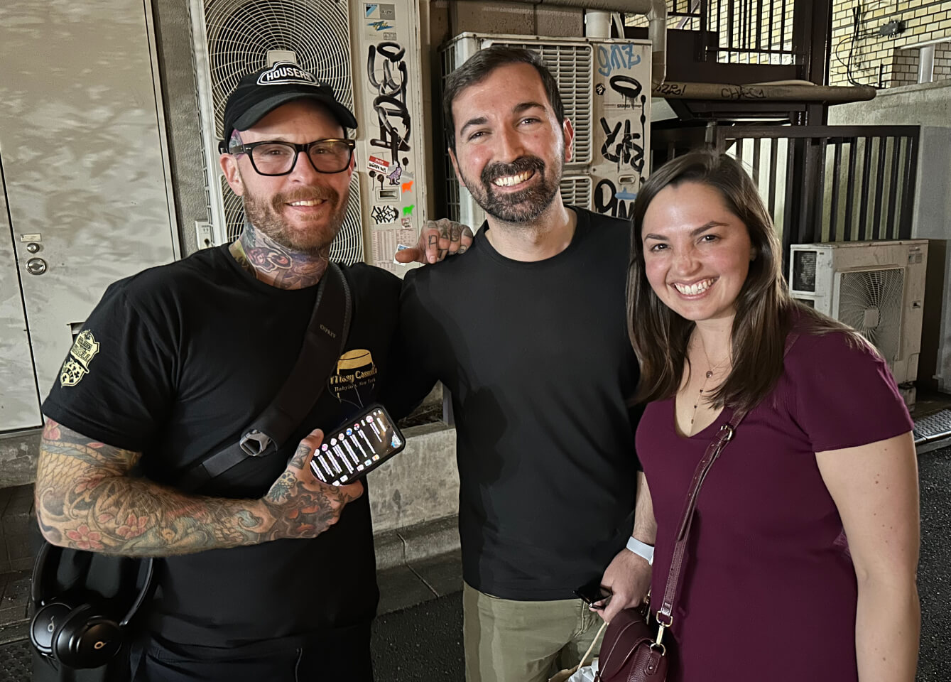 En tatuerad amerikansk man med glasögon och keps, en amerikansk man med mörkt hår och skägg och en amerikansk mörkhårig kvinna med vinröd klänning. Alla ler.