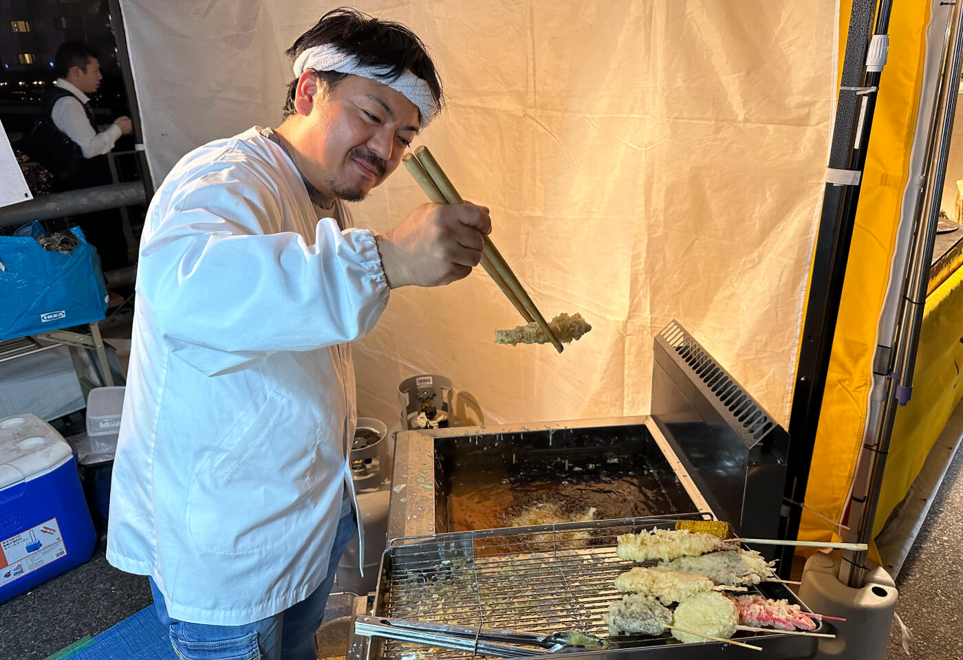 En kock håller i en bit tempura ovanför kokande olja med två tjocka ätpinnar. Han ser nöjd ut.