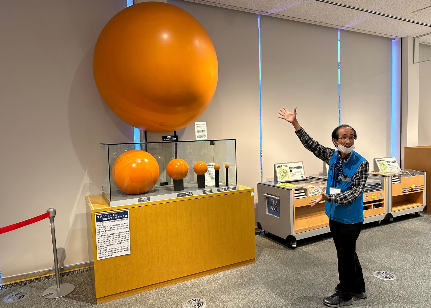 Guide på museum visar sex orangea klot som symboliserar energimängder vid jordbävningar. Ett klort är klart större än de andra.