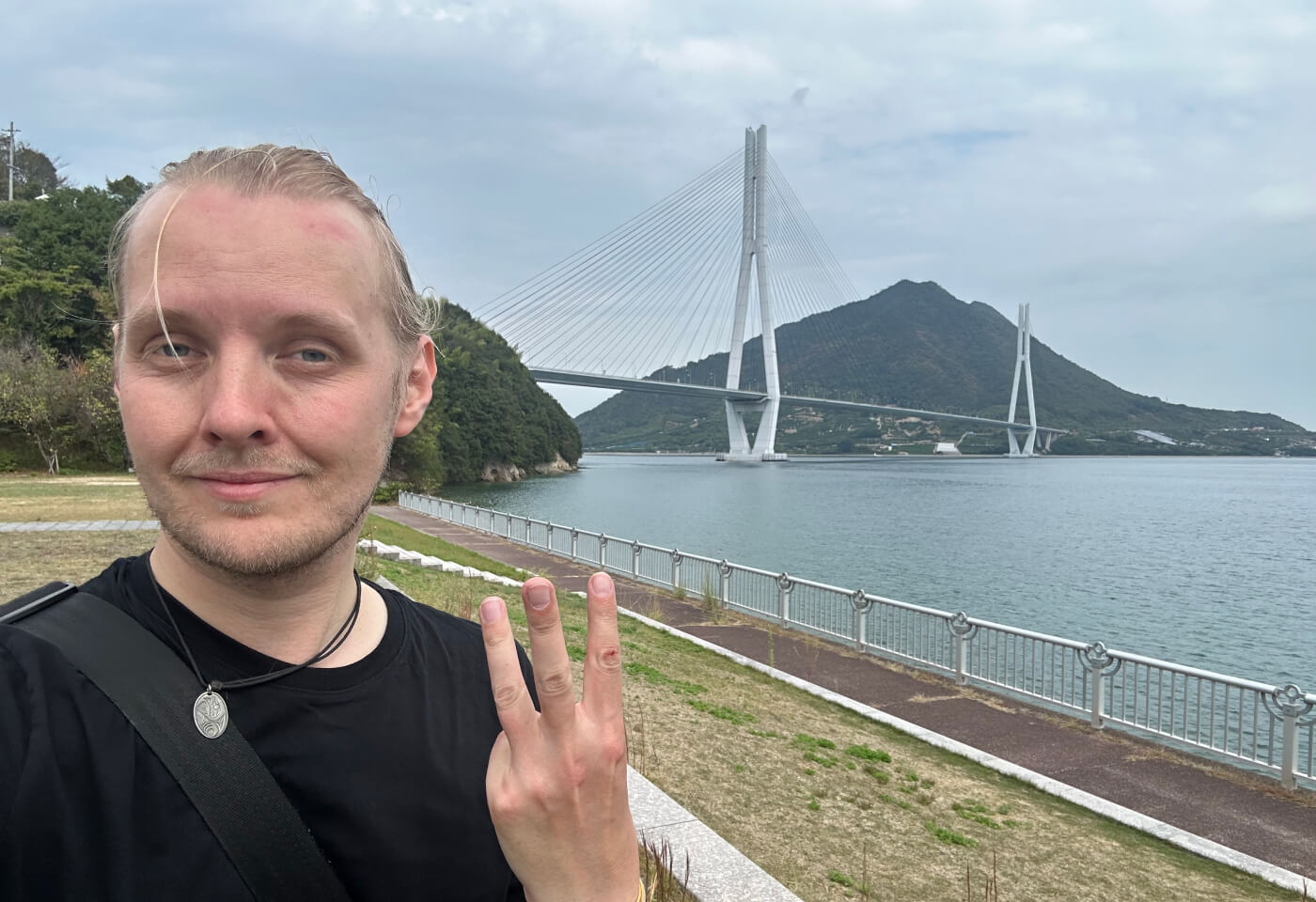 Selfie på mig med uppsatt hår och hållande tre fingrar i luften. Bakom mig en lång vit hängbro över vatten mellan två öar.